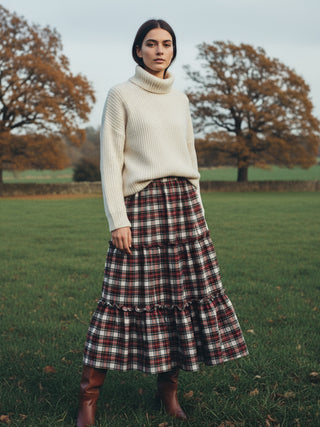 Woman wearing a cream sweater and plaid skirt standing in a field with trees in the background