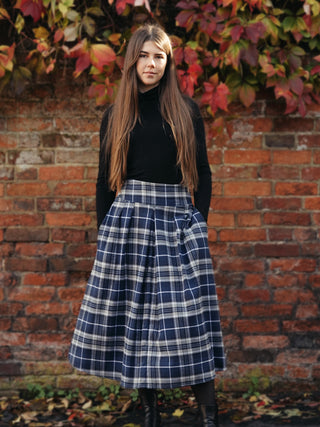 Woman wearing a black turtleneck and plaid skirt standing against a brick wall with autumn leaves.