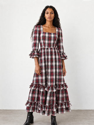 A woman modeling a long-sleeved plaid maxi dress with a square neckline, ruffled cuffs, and tiered ruffles at the hem, paired with black boots against a plain white studio background.