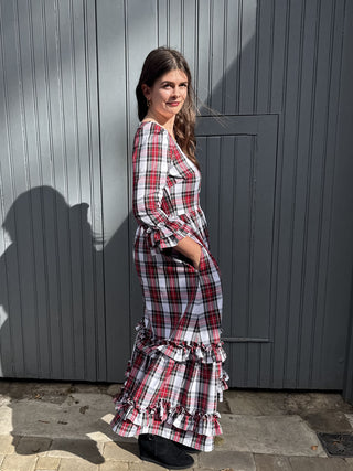 A woman wearing a red, white, and black plaid maxi dress with ruffled details, standing in front of a gray corrugated metal wall.