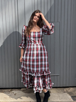 A woman wearing a red, white, and black plaid midi dress with puff sleeves and a tiered ruffle hem, standing in front of a gray corrugated metal wall.