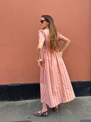 A woman wearing a short-sleeve midi dress with vertical orange and white stripes, standing against a textured terracotta wall.