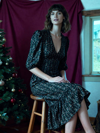 Woman in a dark floral dress sitting on a stool next to a decorated Christmas tree against a red curtain.