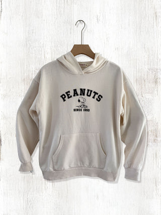 Beige hoodie with 'Peanuts' logo on a wooden hanger against a light wooden background