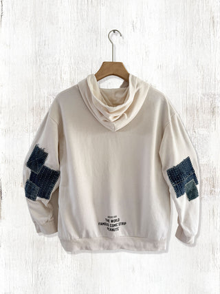 Beige hoodie with denim patch details on a wooden hanger against a white wooden background