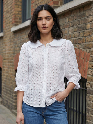 Woman wearing a white blouse with a collar and blue jeans standing against a brick wall.