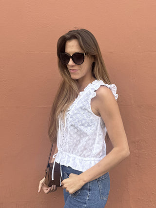 A woman wearing a white sleeveless lace top with ruffle details, blue jeans, and dark sunglasses, standing against a textured terracotta wall and holding a small brown crossbody bag.