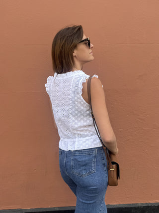 A woman wearing a white sleeveless lace top and blue jeans stands in profile against a textured terracotta wall, carrying a brown crossbody bag.
