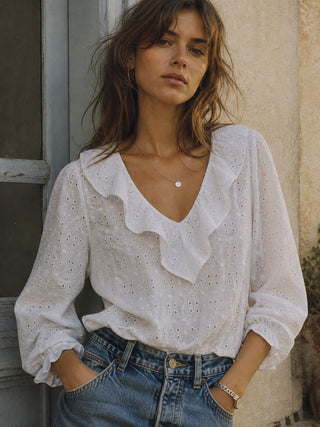 Woman wearing a white blouse with ruffled details and blue jeans, standing against a stone wall.