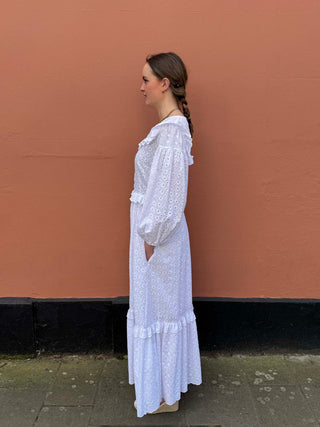 White cotton broderie anglaise dress with button front, gathered puff sleeves and ruffle trims, worn by model against a terracotta wall – handmade to order in the UK by The Well Worn