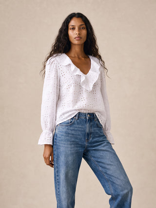 A woman modeling a white eyelet blouse with a ruffled V-neckline and long sleeves, paired with blue jeans, against a plain beige studio background.